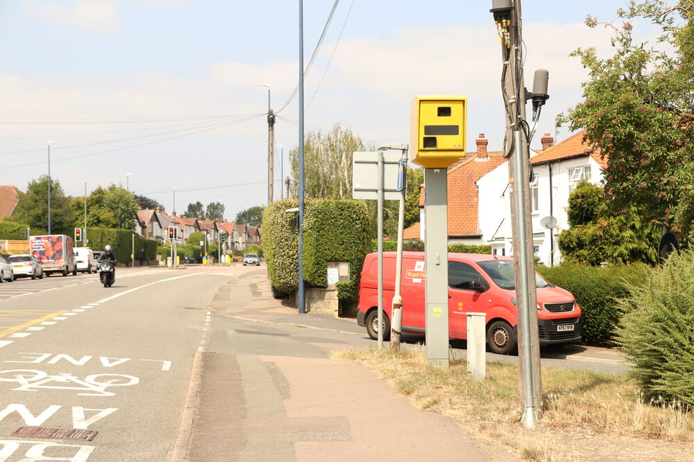 A speed camera partially concealed behind roadside furniture and a hedge — hidden speed traps are still legal