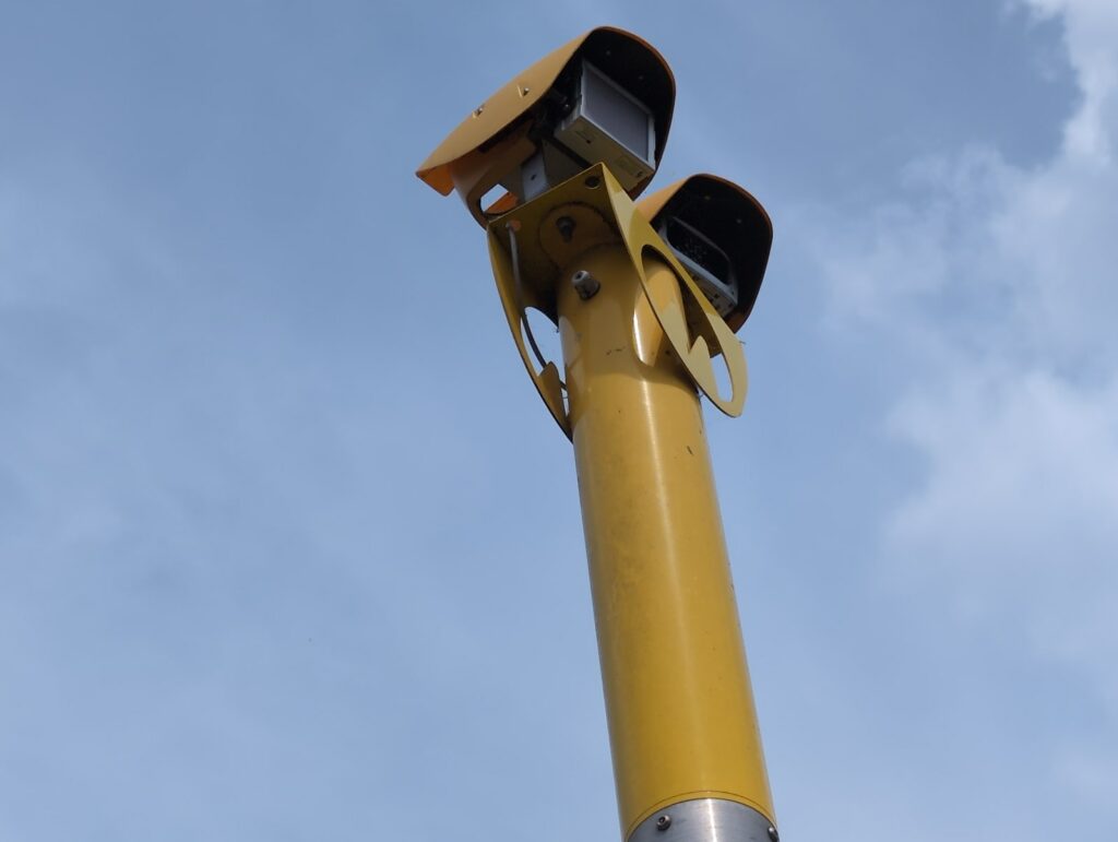 A twin-headed yellow speed camera up close against the sky — one flash can mean six points on your licence