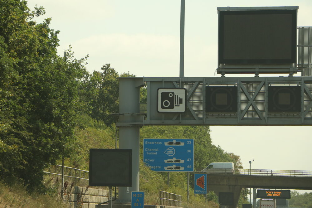 A smart motorway gantry above flowing traffic, where sudden variable limit changes cause hard braking